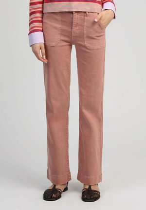 Femme portant un pantalon droit couleur rose, un haut rayé à manches longues et des sandales plates noires tissées, debout avec une main dans la poche.