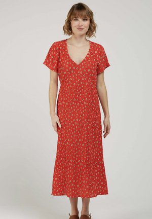 Femme portant une robe midi rouge à fleurs avec des manches courtes et un décolleté en V, debout devant un fond clair uni, faisant face à l'avant.