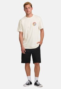 Cream short-sleeve t-shirt with a round logo on the left chest, paired with black shorts. Cotton material, relaxed fit, minimal design.