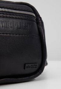 Musta nahkakotelo, jossa on teksturoitu pinta, hopeinen vetoketjukiinnitys ja kohokuvioitu "MORE FLIGHT" -yksityiskohta. Varustettu Alpha Industries -logomerkillä.