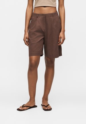 Person iført knælange brune shorts, beige crop top og sorte flip-flops, der står på en ensfarvet baggrund.