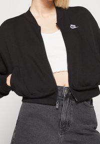 Černá bomber bunda na zip s krátkým střihem. Obsahuje bílé logo Nike, žebrované manžety a přední kapsy. Nositá přes bílý top.