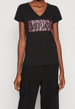 Frau trägt ein schwarzes V-Ausschnitt-T-Shirt mit glänzendem "LIU JO"-Logo und schwarze Cargohose vor einem einfarbigen hellen Hintergrund.