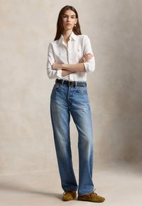 Mujer con camisa blanca abotonada, vaqueros azules, cinturón negro y mocasines marrones, de pie con los brazos cruzados frente a un fondo liso.