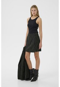 Schwarzes Tanktop kombiniert mit einem dunkel gemusterten Rock mit vertikalen Linien und einem Knopfdetail; das Modell hält einen passenden Mantel; trägt schwarze Stiefel.
