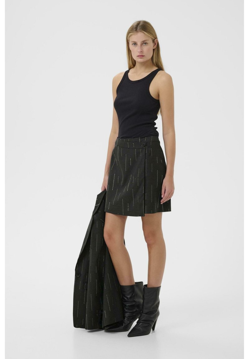 Top nero senza maniche abbinato a una gonna scura con motivo a righe verticali e dettaglio dei bottoni; la modella tiene un cappotto coordinato; indossa stivali neri.