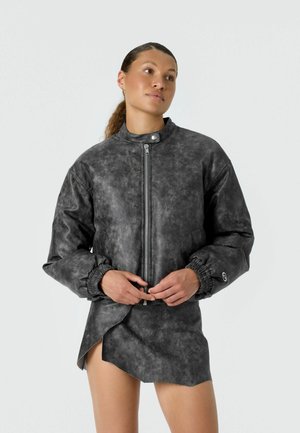 Femme portant une veste zippée texturée gris foncé avec des poignets élastiques et une mini-jupe asymétrique assortie, debout, les mains jointes.