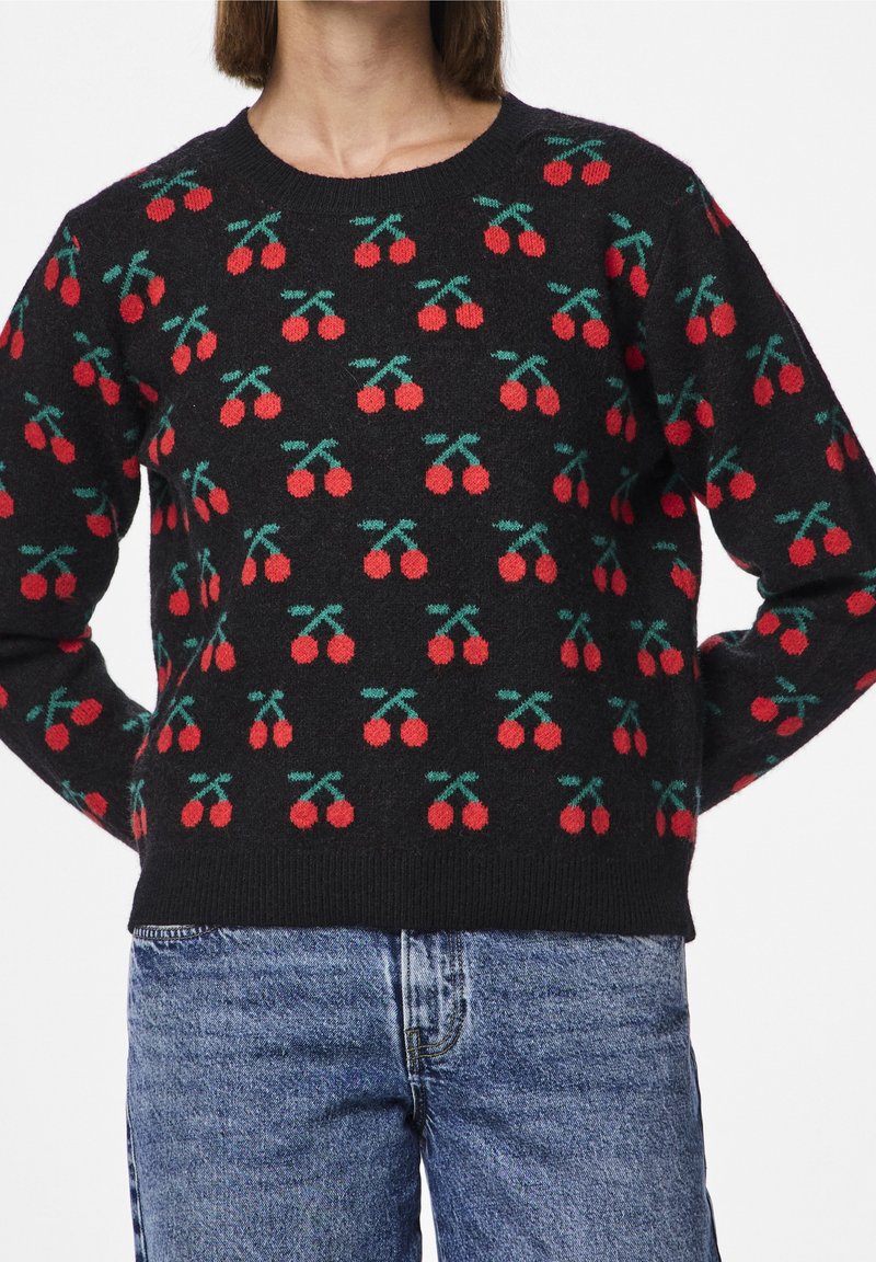 Pull noir avec motifs de cerises rouges porté avec un jean bleu, visible du cou aux hanches, visage de la personne non visible.