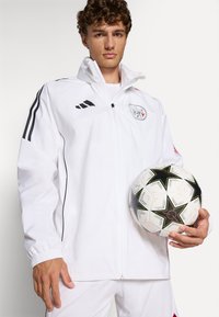 Hvid jakke med lynlås, sorte striber og logo, parret med en mønstret fodbold med sorte stjerner på en hvid baggrund.