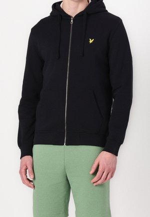 Mann trägt schwarzen Kapuzenpullover mit Reißverschluss und gelbem Vogel-Logo, kombiniert mit hellgrünen Hosen, steht vor weißem Hintergrund.