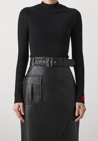 Eine Frau trägt ein schwarzes, eng anliegendes Langarmoberteil und einen figurbetonten, schmalen schwarzen Lederrock mit einem Gürtel und seitlichen Taschen.