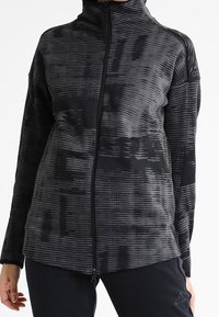 Veste zippée noire avec un motif texturé à rayures. Présente un col haut, des manches longues et un logo Adidas sur l'épaule.