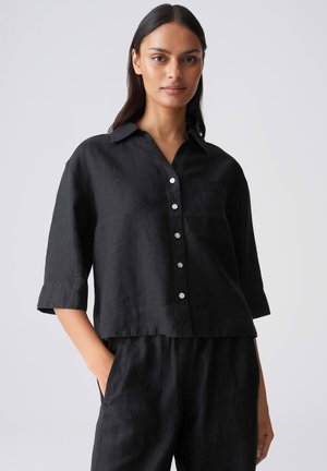 Femme portant une chemise noire ample à boutons avec des manches mi-longues et un pantalon noir assorti, debout avec une main dans la poche.