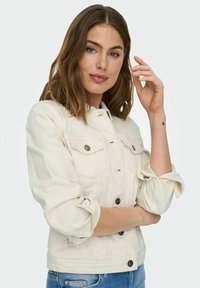Light beige denim jacket with a fitted design, button closures, two chest pockets, and rolled-up sleeves, paired with blue jeans.