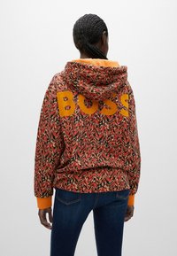 Sudadera con capucha de patrón zigzag en naranja y rojo, con "BOSS" en letras amarillas negritas en la parte posterior y puños naranjas en contraste.