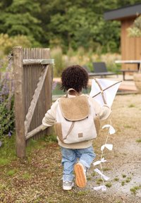 Sac à dos en fausse fourrure beige et blanche avec un rabat, une poche latérale et une étiquette de logo. Enfant tenant un cerf-volant avec un tissu blanc et un cadre en bois, marchant en extérieur.
