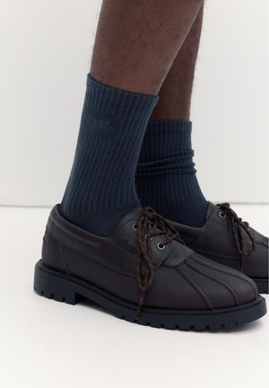 Pantofi maro închis din piele cu șireturi, purtați cu șosete bleumarin cu dungi, pe o persoană cu pielea închisă la culoare, stând pe un podea deschisă la culoare.