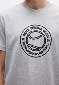 Pilka trumpomis rankovėmis marškinėliai su juodu apskritimo formos atspaudu, vaizduojančiu teniso kamuoliuką ir tekstu: "BOSS TENNIS CLUB 1997 ESTABLISHED SPORTS BRAND."