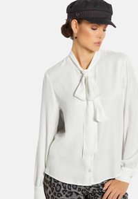 Weißes Blusenshirt mit einer Schleife am Kragen, langen Ärmeln und Knopfverschluss, kombiniert mit schwarzen und grauen Leopardenmuster-Hosen und einer schwarzen Kappe.