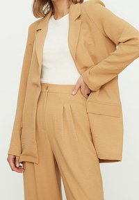 Blazer de tela arrugada beige con un solo botón y un bolsillo, combinado con pantalones plisados a juego y una blusa blanca.