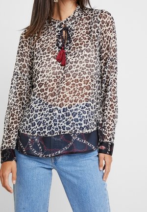 Kobieta nosząca przezroczystą bluzkę w panterkę z czarną i czerwoną frędzlą, nałożoną na wzorzysty czarny top i jasnoniebieskie jeansy.