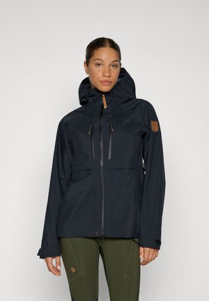 Marineblauwe waterdichte jas met een hoge kraag, twee voorzakken met rits, verstelbare manchetten en een logo-patch op de mouw. Glad fabric textuur.