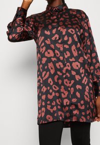 Chemise en satin noir avec un imprimé léopard rouge, manches longues, fermeture éclair à boutons à l'avant et un ourlet légèrement allongé. Texture lisse, coupe décontractée.