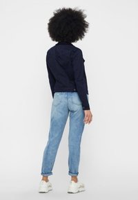 Giacca di denim blu navy con maniche lunghe, chiusura a bottoni e design cropped, abbinata a jeans azzurri e sneakers bianche.