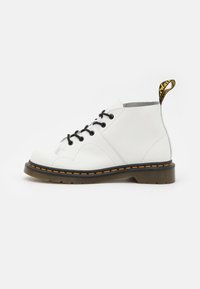 Botas de tobillo de cuero blanco con cordones negros, tirador amarillo y negro, y un detalle de costura naranja contrastante a lo largo de la suela. Suela de goma.