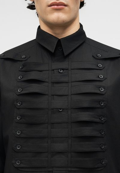 Primo piano di una persona che indossa una maglietta nera con cinturini decorativi paralleli con bottoni sul petto e sulle spalle.