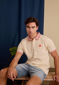 Polohemd in lichtbeige met bladdessin, gestreepte rode kraag en mouwen. Draag het met lichtblauwe denimshorts, zittend op een houten bank.