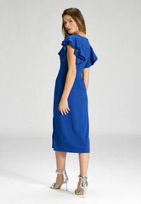 Robe bleue avec des manches volantées, corsage ajusté, jupe mi-longue et fente sur le côté. Associée à des sandales à talons hauts argentées.