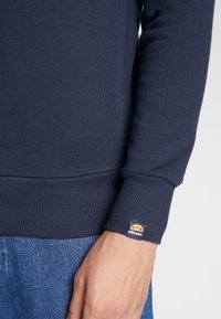 Felpa blu navy realizzata in tessuto a costine, con colletto e polsini aderenti. Presenta un piccolo tag con il logo ellesse sul polsino sinistro.