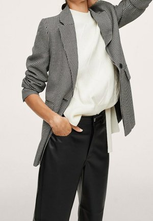 Persoon draagt zwarte leren broek, witte gebreide trui en zwart-wit pied-de-pouled-blazer met opgestroopte mouwen, hand in de zak.