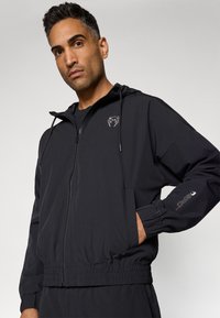 Sort zip-up jakke med hætte, lavet af letvægtsstof. Har elastiske manchetter og talje, samt et logo på brystet og ærmet.