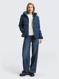 Vrouw staand, gekleed in een blauw pufferjack, witte trui, wijde jeans en zwarte laarzen tegen een effen lichte achtergrond.