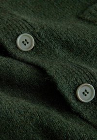 Donkergroene wollen stof met een zachte, textuurrijke oppervlakte. Voorzien van twee lichtgekleurde knopen, elk met vier gaten om vast te maken.