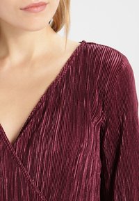 Burgundy pleated top with a deep V-neckline and textured fabric, featuring vertical grooves for added detail.