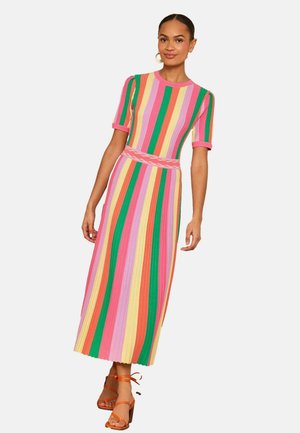 Moteris vilki spalvotą midi suknelę su vertikaliomis juostelėmis, trumpomis rankovėmis ir oranžinius sandalėlius su dirželiais, stovi prieš baltą foną.