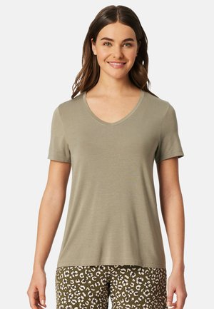 Jonge vrouw met lang bruin haar, gekleed in een licht olijfgroen V-hals t-shirt met korte mouwen en een luipaardprint broek, glimlachend.