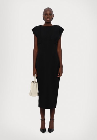 Black fitted dress with cap sleeves, mid-calf length, structured design. Model holds a small cream handbag and wears black pointed heels.