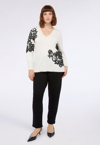 Cremfarbener Pullover mit schwarzer Spitzenverzierung am linken Ärmel und an der Seite; V-Ausschnitt, lange Ärmel, kombiniert mit schwarzen Hosen und spitzen Absätzen.