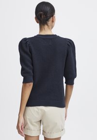 Marineblauer Strickpullover mit strukturiertem Muster, gepufften kurzen Ärmeln und gerippten Bündchen. Kombiniert mit hellbeigen Shorts.