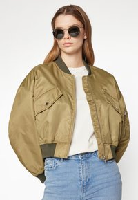 Jachetă bomber de culoare verde măsline, confecționată dintr-un material lucios, având un tiv striat, două buzunare frontale și închidere cu fermoar. Purta tătipă peste un top deschis și blugi.