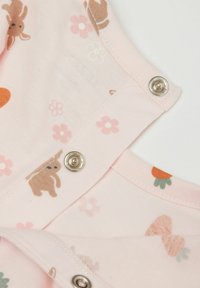 Růžová látka s malými vzory králíčků a květin, s kovovými patentkami na rameni. Měkká textura, hravé designové prvky.