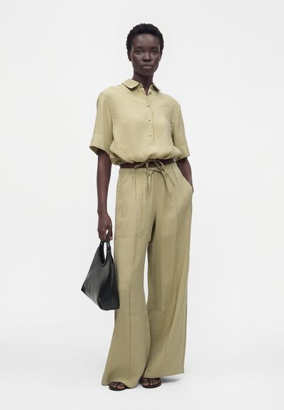 Femme portant une chemise ample à manches courtes couleur olive claire et un pantalon ample, tenant un sac à main noir, debout sur un fond blanc.