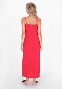 Abito maxi rosso plissettato con spalline sottili e scollatura volant. Presenta un design fluido, indossato con sandali piatti. Vista posteriore mostrata.