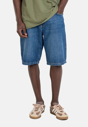 Hombre vistiendo shorts de mezclilla azul, camisa verde oliva y zapatillas beige y marrón con rayas blancas, de pie con una mano en el bolsillo.