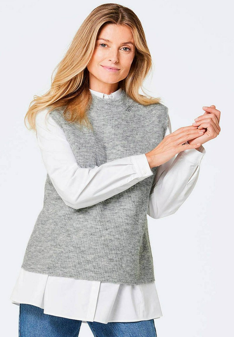 Grau gestrickter ärmelloser Pullover über einem weißen Hemd, mit hohem Halsabschluss und gerippter Textur. Darunter werden hellblaue Jeans getragen.