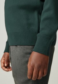 Dunkelgrüner Strickpullover mit gerippten Bündchen und Saum. Verfügt über ein dezentes "Clarks"-Logo auf der unteren Vorderseite. Kombiniert mit strukturierten grauen Hosen.